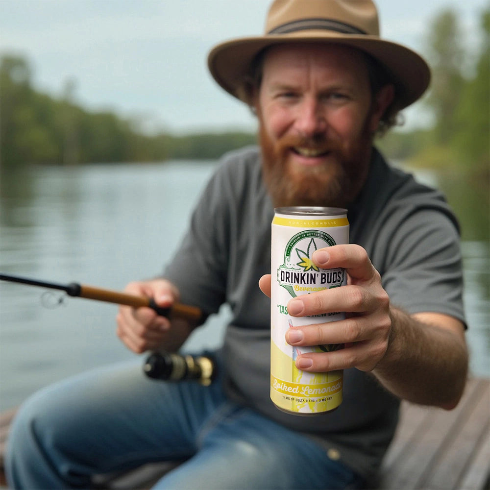 A bearded man in a hat holds out a can of Drinkin’ Buds Spiked Lemonade while fishing on a calm lake, smiling as he enjoys the outdoors.
