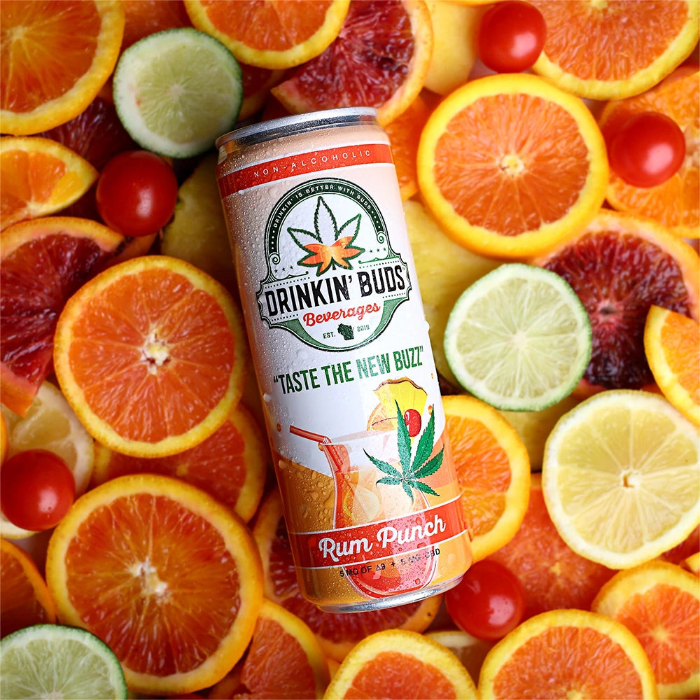 A chilled can of Drinkin’ Buds Rum Punch resting on a vibrant bed of sliced oranges, limes, lemons, and cherry tomatoes, emphasizing its fruity and refreshing flavors.