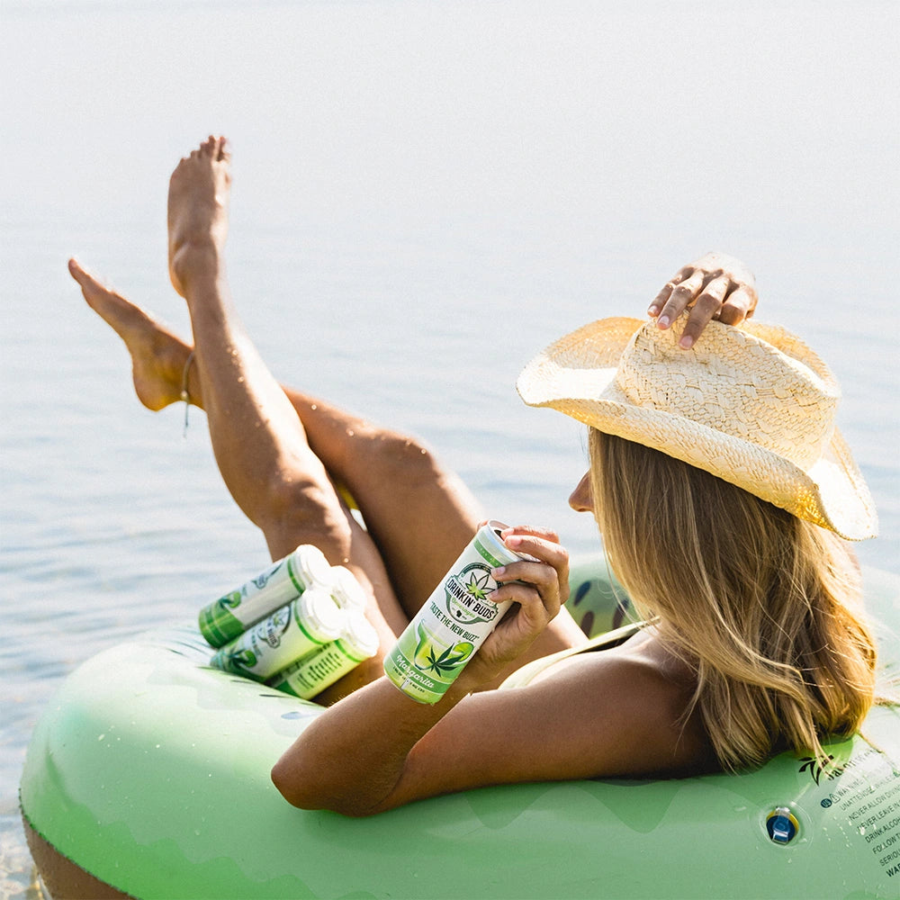 
                      
                        A woman in a straw hat lounges on a green inflatable tube in the water, holding a can of Drinkin’ Buds Margarita while a four-pack rests beside her.
                      
                    