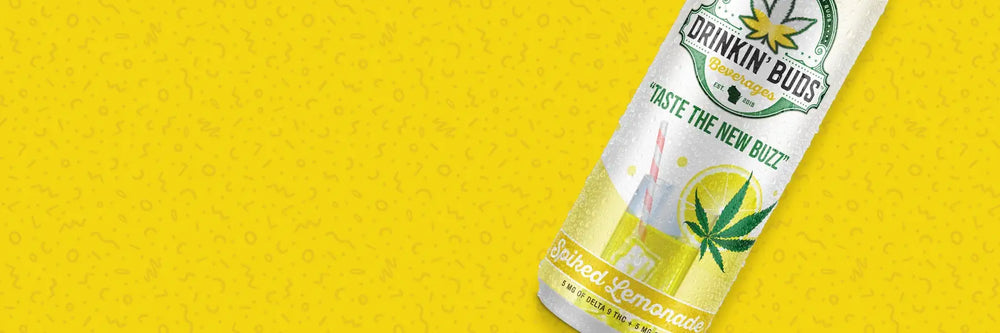 A bright yellow promotional banner for Drinkin' Buds Beverages featuring a Spiked Lemonade-flavored can. The can shows an illustration of a lemonade glass with a red-striped straw, a lemon slice, and a cannabis leaf garnish. The tagline reads, "Taste the New Buzz," with the label highlighting 5 mg of Delta-9 THC and 5 mg of CBD, promoting it as a refreshing, cannabis-infused beverage perfect for summer vibes.