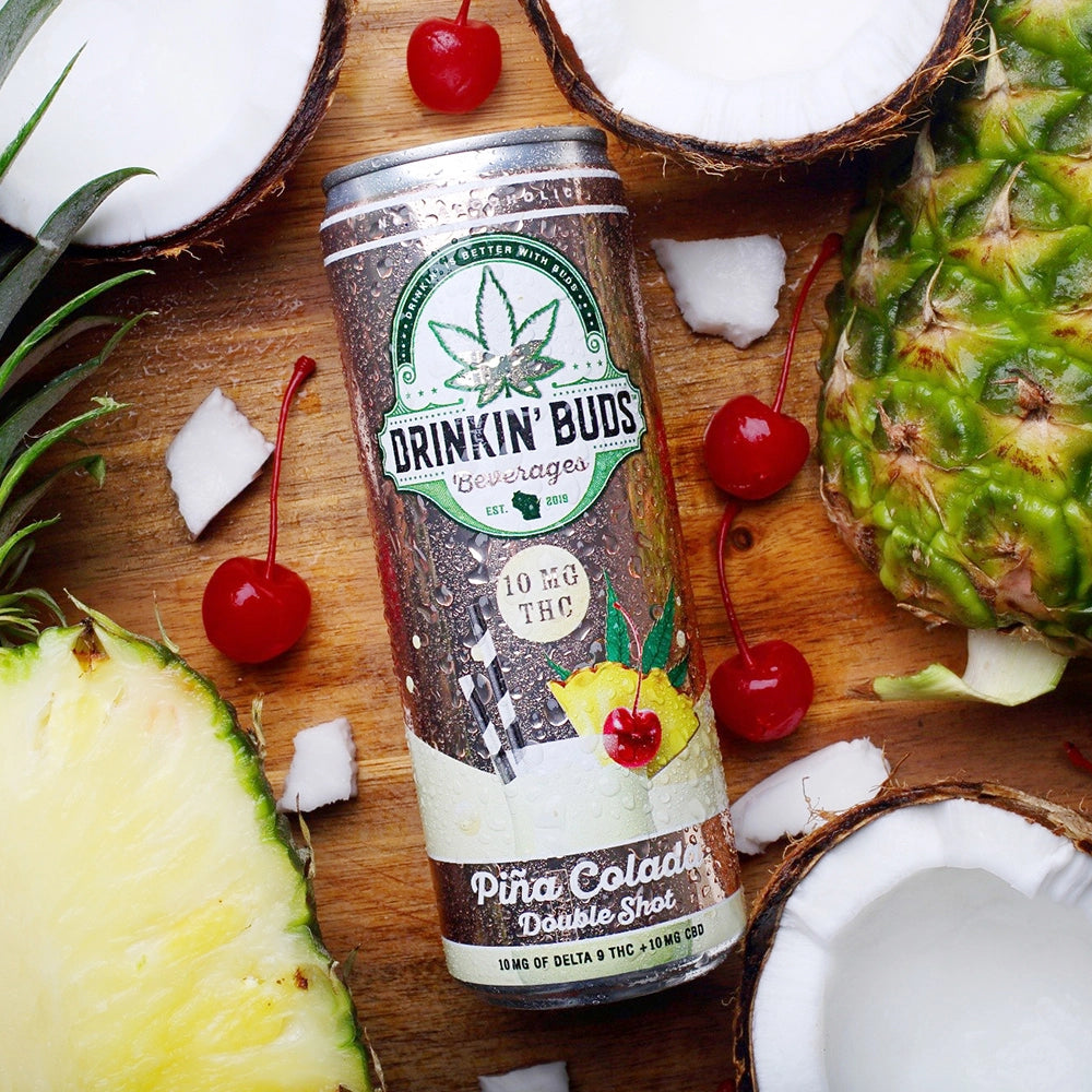 
                      
                        A chilled can of Drinkin' Buds Piña Colada Double Shot is surrounded by fresh pineapples, coconuts, and cherries on a wooden surface.
                      
                    
