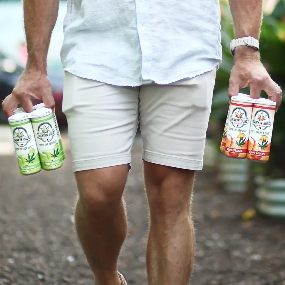 This image captures a person casually walking while carrying two four-packs of "Drinkin' Buds" canned beverages. One set features margarita-flavored cans with green accents, and the other set showcases rum punch-flavored cans with orange-red tones. The individual is dressed in light-colored shorts and a casual shirt, emphasizing a laid-back vibe, likely heading to or from a gathering. The focus on the hands holding the beverages suggests portability and convenience.