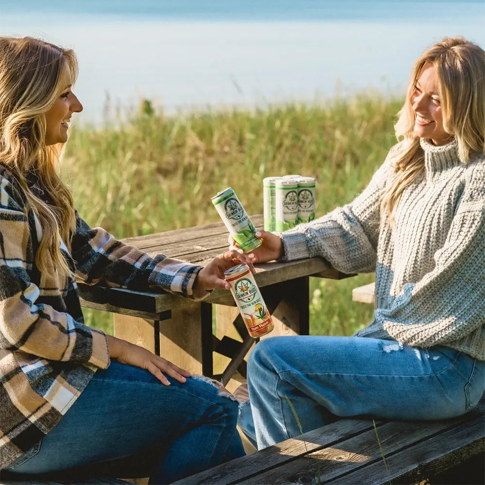 This image captures two women sitting at a rustic wooden picnic table near a grassy lakeside area, enjoying cans of "Drinkin' Buds" beverages. One holds a margarita-flavored can, while the other offers a rum punch-flavored can, highlighting a moment of relaxation and connection. Both women are dressed warmly in cozy sweaters, suggesting a cool, outdoor setting perfect for a casual drink by the water. Multiple cans are stacked on the table, emphasizing the beverage’s social and refreshing appeal.