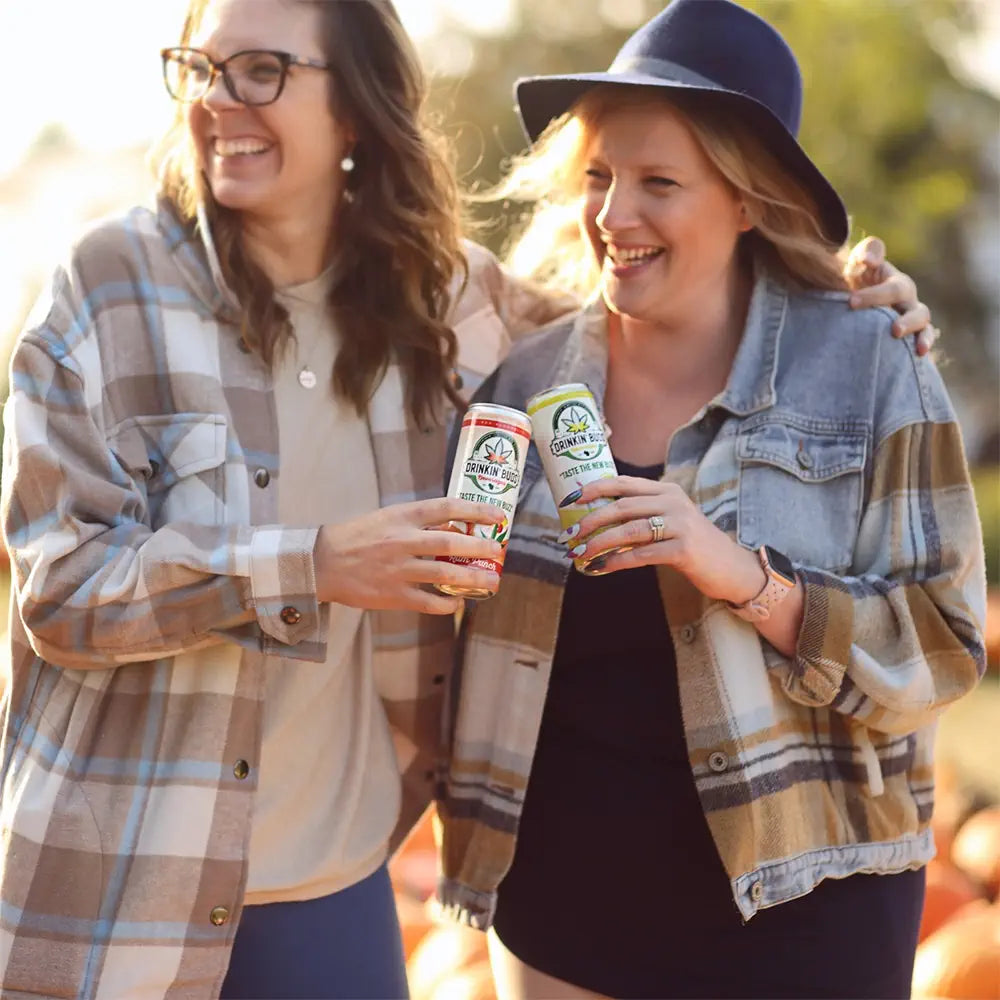 This image shows two smiling women enjoying cans of "Drinkin' Buds" beverages outdoors, embracing a cheerful, social moment. One holds a rum punch-flavored can, and the other holds a margarita-flavored one, both with the tagline "Taste the New Buzz" and cannabis-themed branding. Dressed in plaid jackets and casual layers, the women appear relaxed and joyful, suggesting a cozy, friendly gathering in cool weather.