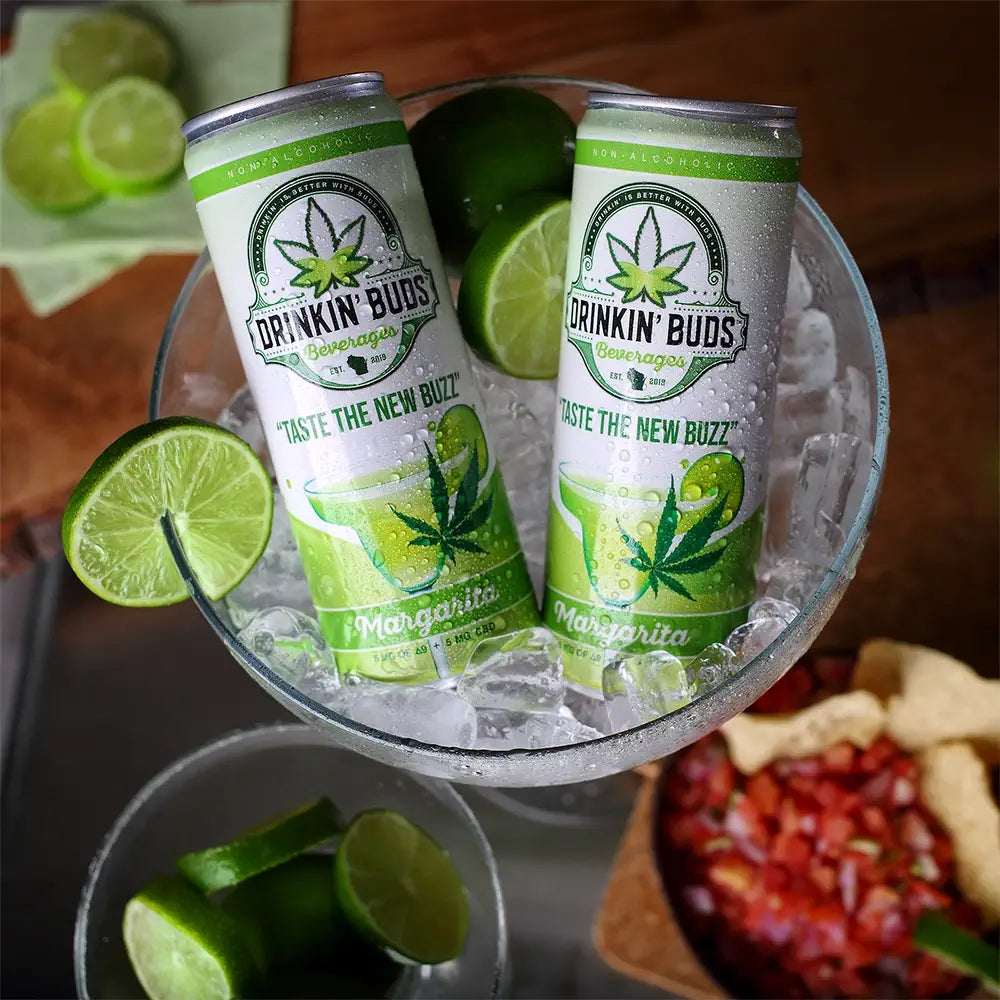 This image showcases two cans of "Drinkin' Buds" margarita-flavored beverages nestled in a large glass filled with ice, surrounded by fresh lime slices. The cans feature the tagline "Taste the New Buzz" along with a cannabis leaf, emphasizing the drink’s non-alcoholic CBD infusion. The vibrant presentation, with salsa and chips in the background, suggests a festive or social gathering atmosphere, perfect for enjoying refreshing, cannabis-infused drinks.