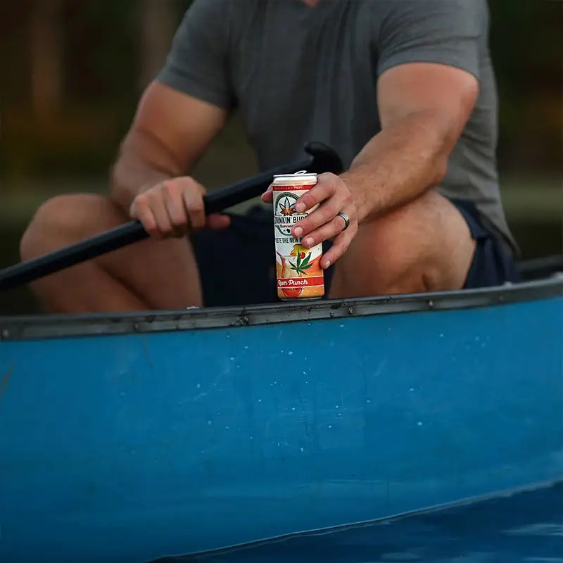 
                      
                        The image shows a person holding a "Drinkin' Buds Beverages" Rum Punch can while seated in a blue canoe, paddle in hand. The scene captures a relaxed, outdoor adventure vibe on the water.
                      
                    